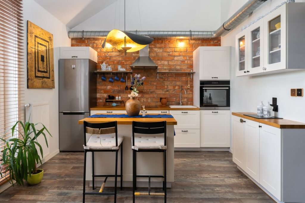 Industrial farmhouse kitchen featuring white shaker cabinets, exposed brick backsplash, wooden countertops, kitchen island, pendant lighting, and stainless steel appliances.